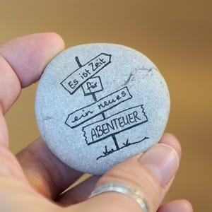 Könnte beinhalten: Ein grauer Stein mit schwarzen Tintenzeichnungen eines Wegweisers mit vier Pfeilen, die in verschiedene Richtungen zeigen. Der Wegweiser lautet "Es ist Zeit fur ein neues Abenteuer", was übersetzt "Es ist Zeit für ein neues Abenteuer" bedeutet.