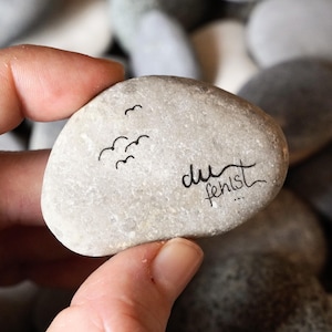 Könnte beinhalten: Ein glatter, grauer Stein mit drei kleinen, schwarzen Vogelsilhouetten. Der Satz "du fehlst..." ist in schwarzer Schrift geschrieben. Der Stein wird in einer Hand gehalten, mit anderen Steinen im Hintergrund.