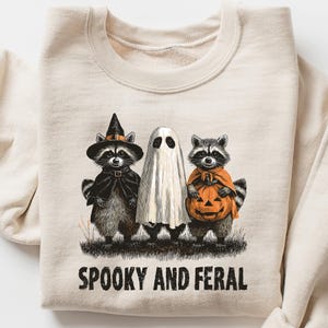 May include: Cream-colored sweatshirt featuring a Halloween-themed graphic. The design includes three raccoons dressed as a witch, a ghost, and one holding a pumpkin. The text "SPOOKY AND FERAL" is printed below the illustration.