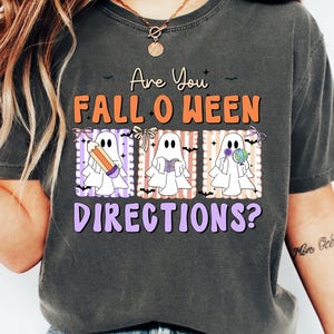 May include: Dark gray t-shirt with the text "Are You Fall O Ween Directions?" in orange and purple. The shirt features three cartoon ghosts holding a pencil, a book, and a lollipop. The shirt is a casual, comfortable style.