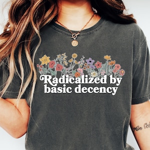 May include: Dark gray t-shirt with the phrase "Radicalized by basic decency" in white script. The shirt features a floral design across the chest with various colorful flowers. The model is wearing blue jeans and white sneakers.