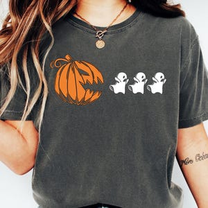 May include: Dark gray t-shirt featuring a Halloween design. An orange pumpkin with a carved face is on the left, and three white ghost figures are to the right. The shirt is a casual style.
