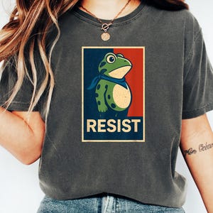 May include: Dark gray t-shirt featuring a frog graphic with a blue bandana and the word "RESIST" in a rectangular design. The frog is green with black spots. The background is red and blue.