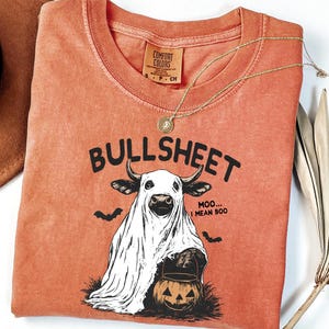 May include: Orange Comfort Colors t-shirt with a Halloween-themed graphic. The graphic features a cow in a ghost costume with the text "BULLSHEET" above and "MOO... I MEAN BOO" below. Includes a gold necklace and a brown hat.