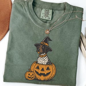 May include: Olive green Comfort Colors t-shirt featuring a Halloween-themed graphic. The graphic depicts a dog wearing a witch hat, sitting inside a carved pumpkin. A smaller pumpkin is next to the larger one. A gold necklace is draped on the shirt.