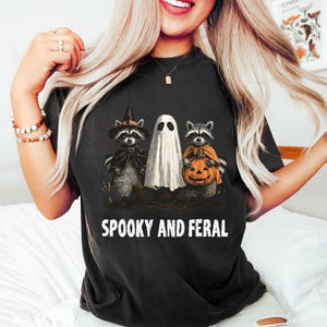 May include: Black t-shirt with a Halloween graphic. The graphic features a ghost and two raccoons dressed in costumes, one with a witch hat and cape, the other holding a jack-o'-lantern. The text "SPOOKY AND FERAL" is printed below the graphic.