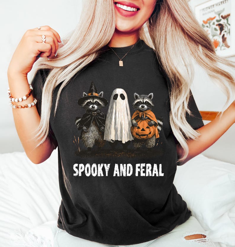 May include: Black t-shirt with a Halloween graphic. The graphic features a ghost and two raccoons dressed in costumes, one with a witch hat and cape, the other holding a jack-o'-lantern. The text "SPOOKY AND FERAL" is printed below the graphic.