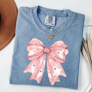 May include: A blue Comfort Colors t-shirt featuring a large pink bow with white ghost illustrations. The bow has a lace-like trim. A gold necklace with a pendant is draped on the shirt. A brown hat and dried plant are in the background.