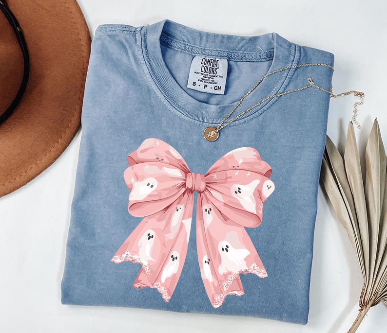 May include: A blue Comfort Colors t-shirt featuring a large pink bow with white ghost illustrations. The bow has a lace-like trim. A gold necklace with a pendant is draped on the shirt. A brown hat and dried plant are in the background.