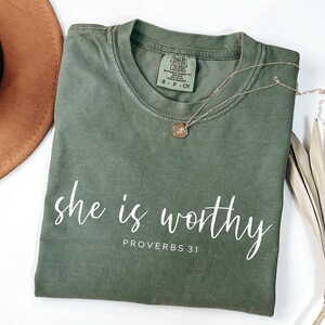 Op de afbeelding: Olijfgroen T-shirt met de tekst "she is worthy" en "PROVERBS 31" in witte letters. Een gouden ketting met een kleine hanger ligt op het shirt. Het shirt is opgevouwen en wordt getoond met een bruine hoed en een gedroogde plant.