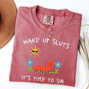 May include: A red-violet t-shirt with the text "WAKE UP SLUTS IT'S TIME TO SIN" in white. The graphic includes a smiling sun wearing sunglasses, a red worm, and blue flowers. A gold necklace is draped across the shirt.