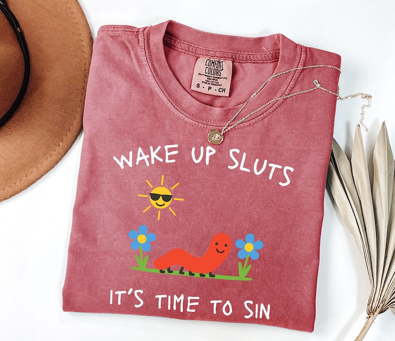 May include: A red-violet t-shirt with the text "WAKE UP SLUTS IT'S TIME TO SIN" in white. The graphic includes a smiling sun wearing sunglasses, a red worm, and blue flowers. A gold necklace is draped across the shirt.