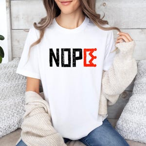 May include: White t-shirt featuring the word "NOPE" in black, distressed block letters. The letter "E" is replaced with a red, stylized crown design. A casual, everyday wear item.