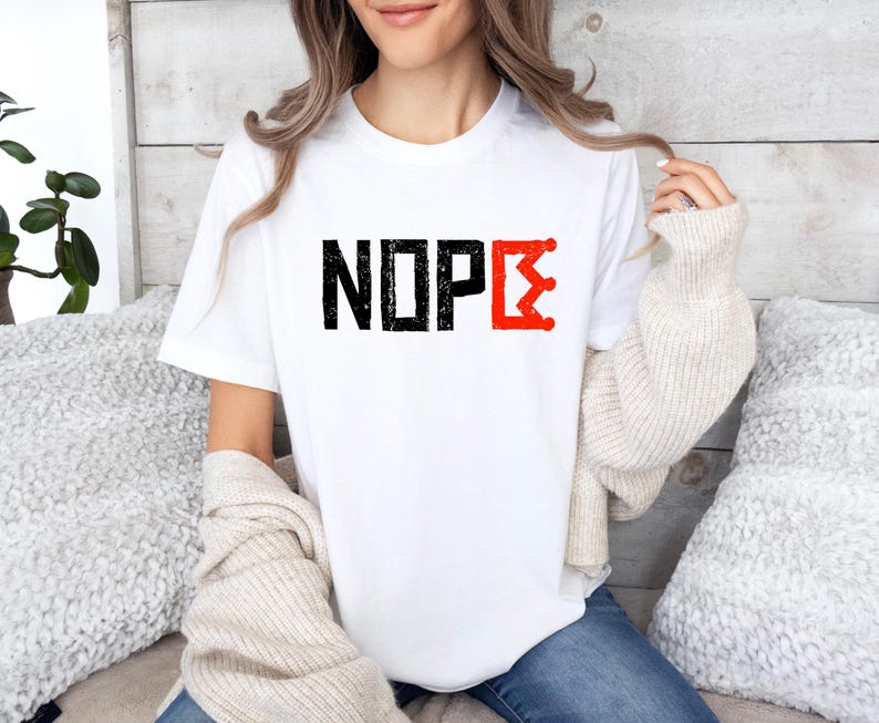 May include: White t-shirt featuring the word "NOPE" in black, distressed block letters. The letter "E" is replaced with a red, stylized crown design. A casual, everyday wear item.