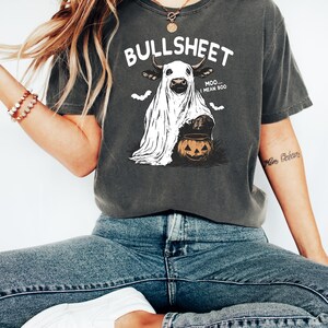 May include: Dark gray t-shirt with a Halloween-themed graphic. The design features a cow dressed as a ghost, holding a jack-o'-lantern, with the text "BULLSHEET...MOO...I MEAN BOO." Two bat silhouettes are above the cow.