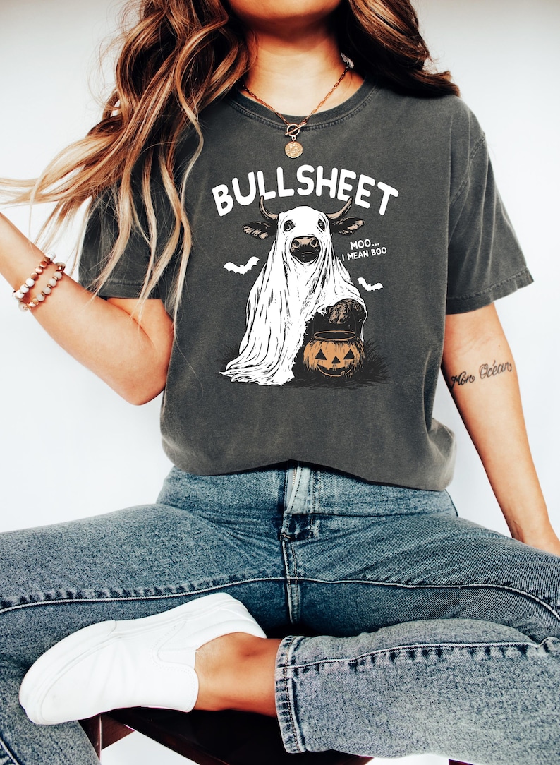 May include: Dark gray t-shirt with a Halloween-themed graphic. The design features a cow dressed as a ghost, holding a jack-o'-lantern, with the text "BULLSHEET...MOO...I MEAN BOO." Two bat silhouettes are above the cow.