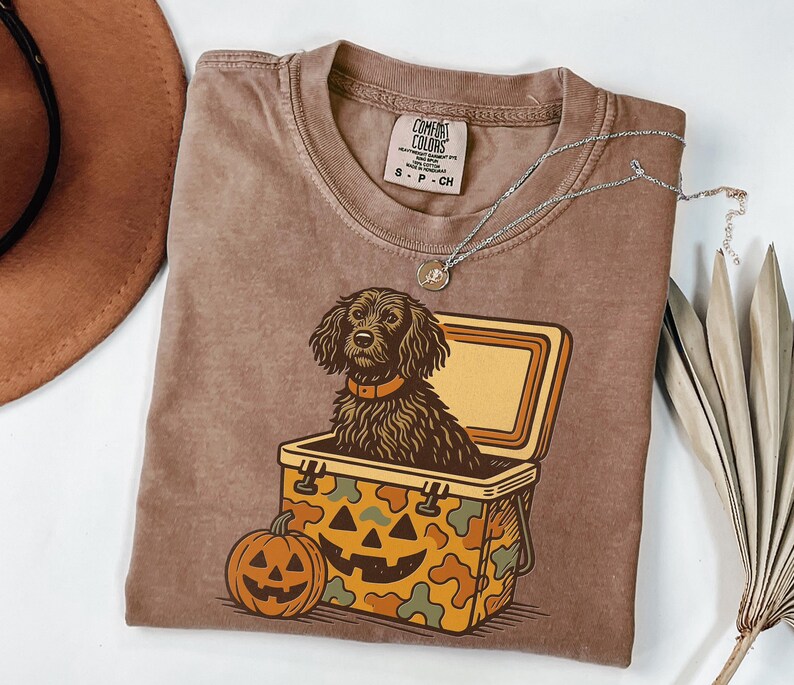 May include: Brown Comfort Colors t-shirt featuring a dog in a jack-o'-lantern cooler. The shirt has a round neck and short sleeves. A silver necklace with a pendant is on the shirt. A brown hat is in the upper left corner.