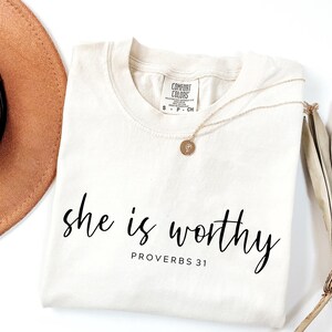 Op de afbeelding: Cr&egrave;mekleurig T-shirt met de woorden "she is worthy" in zwart script, met "PROVERBS 31" eronder. Een gouden ketting met een hanger en een bruine hoed zijn zichtbaar.