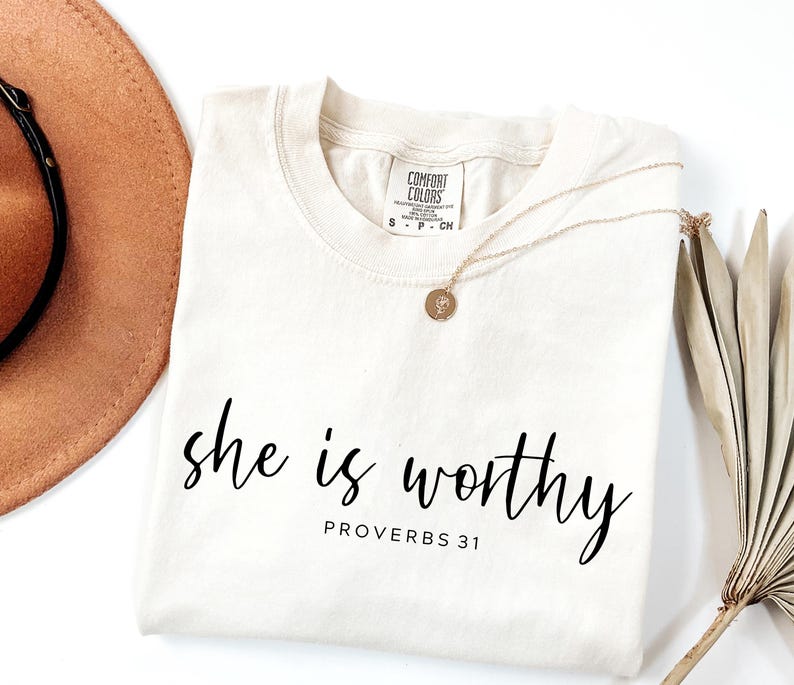 Op de afbeelding: Cr&egrave;mekleurig T-shirt met de woorden "she is worthy" in zwart script, met "PROVERBS 31" eronder. Een gouden ketting met een hanger en een bruine hoed zijn zichtbaar.