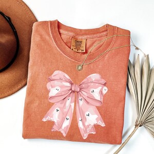 May include: A coral-colored Comfort Colors sweatshirt featuring a large pink bow with white ghost illustrations. A gold necklace with a pendant is draped on the shirt. A brown hat is visible in the upper left corner.