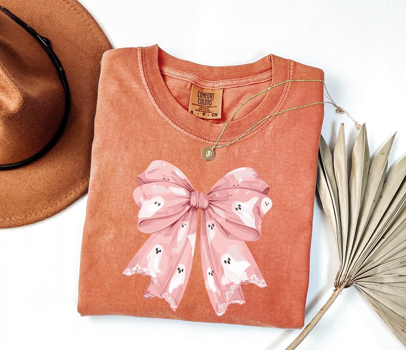 May include: A coral-colored Comfort Colors sweatshirt featuring a large pink bow with white ghost illustrations. A gold necklace with a pendant is draped on the shirt. A brown hat is visible in the upper left corner.