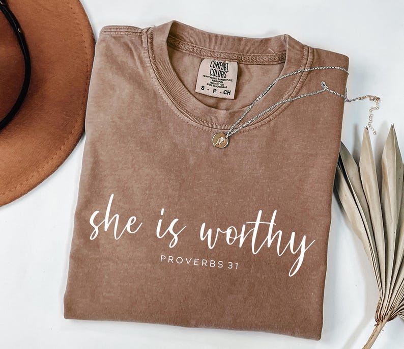 Op de afbeelding: Een bruin Comfort Colors T-shirt met de tekst "she is worthy" en "Proverbs 31" in witte letters. Een zilveren ketting met een kleine hanger ligt op het shirt. Een bruine hoed en gedroogde bladeren zijn op de achtergrond.