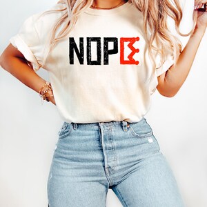 May include: A cream-coloured t-shirt featuring the word "NOPE" in black block letters and a red crown graphic. The shirt is worn with light blue denim jeans.