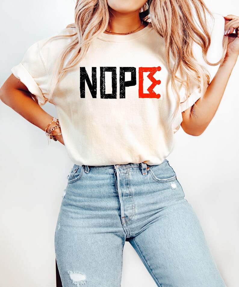 May include: A cream-coloured t-shirt featuring the word "NOPE" in black block letters and a red crown graphic. The shirt is worn with light blue denim jeans.