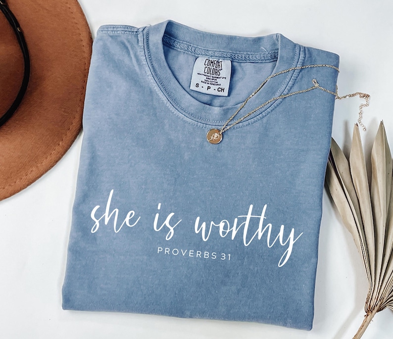 Op de afbeelding: Een blauw t-shirt met de woorden "she is worthy" en "PROVERBS 31" in het wit gedrukt. Een gouden ketting met een hanger ligt op het shirt. Een bruine hoed en een gedroogde plant staan op de achtergrond.