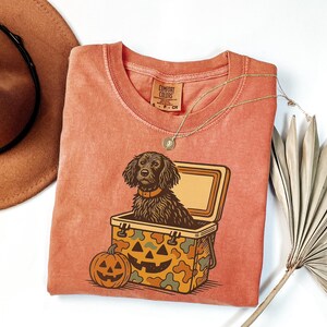 May include: A coral-colored t-shirt featuring a graphic of a dog in a cooler with a jack-o'-lantern. The dog has brown fur and a collar. The cooler has a camouflage pattern. A brown hat and a gold necklace are also visible.