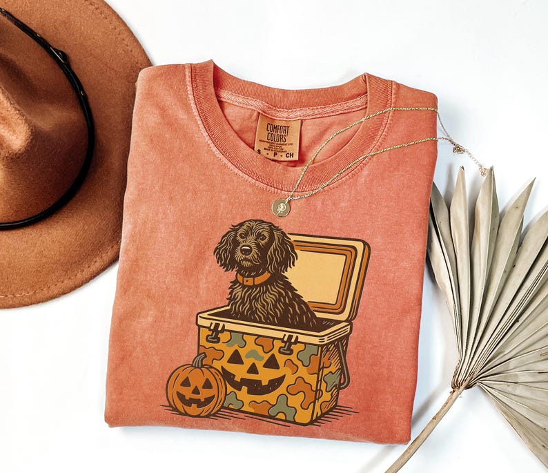 May include: A coral-colored t-shirt featuring a graphic of a dog in a cooler with a jack-o'-lantern. The dog has brown fur and a collar. The cooler has a camouflage pattern. A brown hat and a gold necklace are also visible.