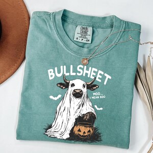 May include: A teal Comfort Colors t-shirt with the text "BULLSHEET" and a graphic of a cow dressed as a ghost holding a jack-o'-lantern. The shirt also has the text "Moo... I mean Boo". A silver necklace with a pendant is on the shirt.