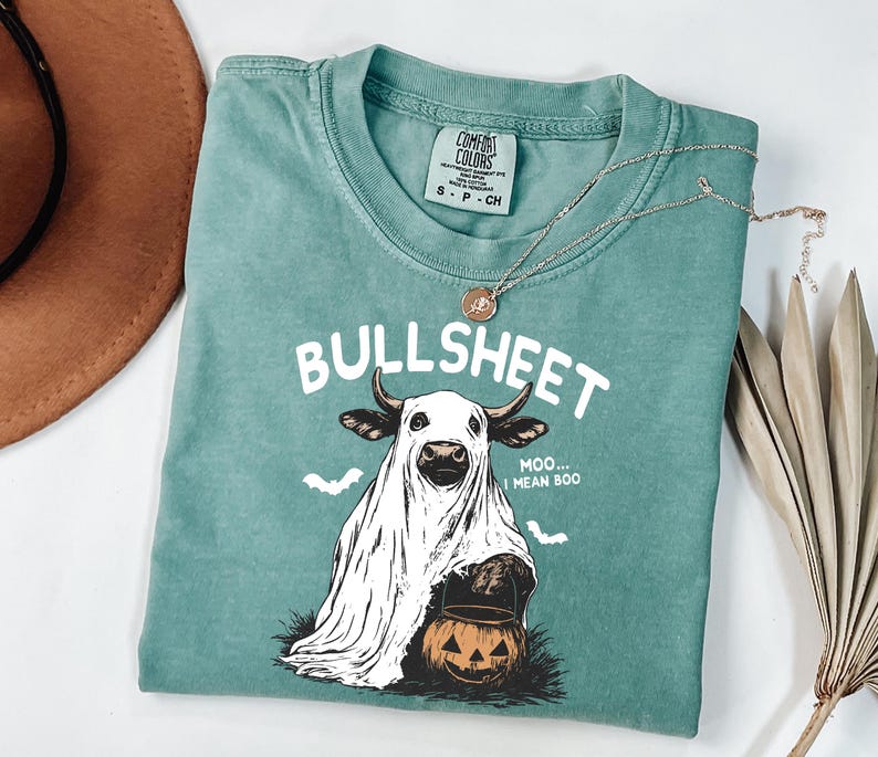 May include: A teal Comfort Colors t-shirt with the text "BULLSHEET" and a graphic of a cow dressed as a ghost holding a jack-o'-lantern. The shirt also has the text "Moo... I mean Boo". A silver necklace with a pendant is on the shirt.