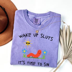 May include: A lilac-colored t-shirt with the text "WAKE UP SLUTS" and "IT'S TIME TO SIN" in black. The shirt features a cartoon sun wearing sunglasses, a smiling orange worm, and blue flowers. A gold necklace is draped over the shirt.