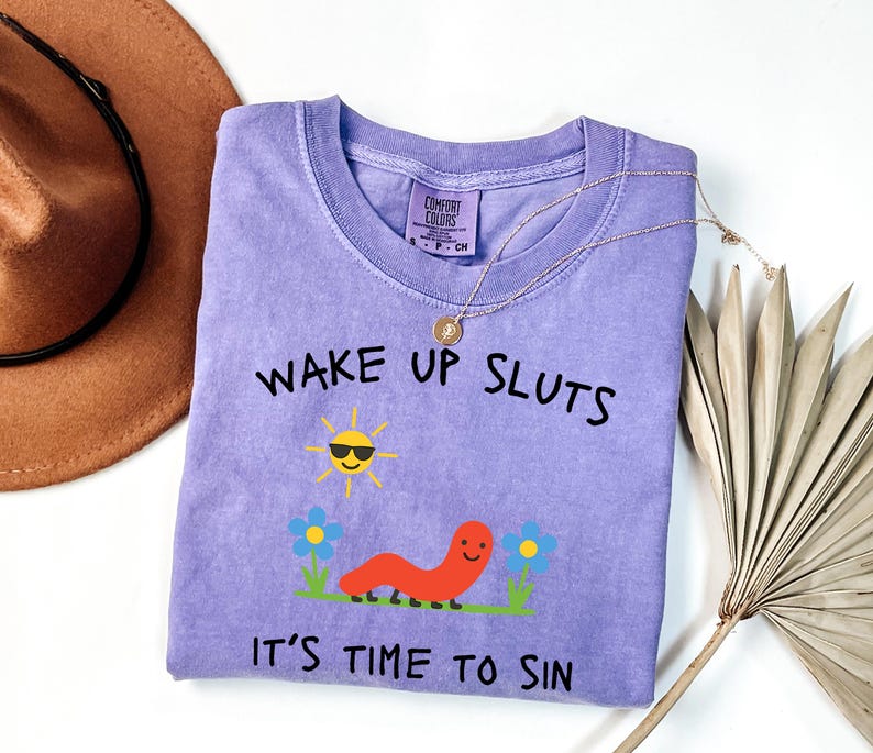 May include: A lilac-colored t-shirt with the text "WAKE UP SLUTS" and "IT'S TIME TO SIN" in black. The shirt features a cartoon sun wearing sunglasses, a smiling orange worm, and blue flowers. A gold necklace is draped over the shirt.