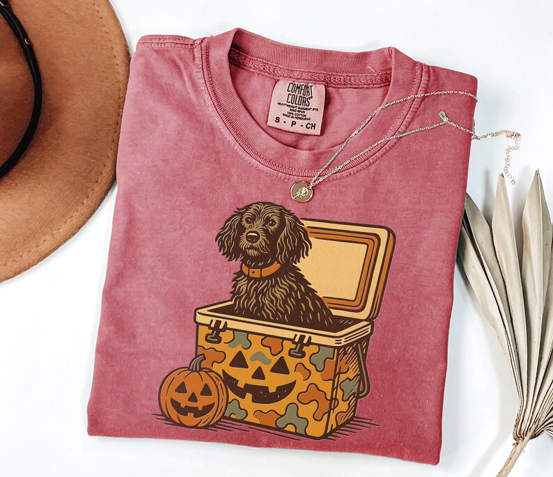 May include: A maroon Comfort Colors t-shirt featuring a dog inside a cooler with a jack-o'-lantern design. The dog has brown fur and is wearing a collar. A pumpkin is next to the cooler. A silver necklace is on the shirt.