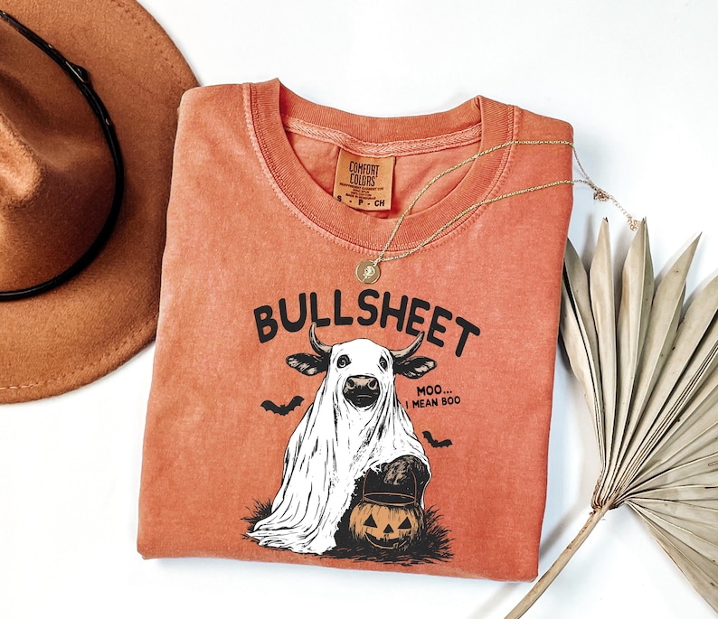 May include: Orange Comfort Colors t-shirt with a Halloween-themed graphic. The graphic features a cow in a ghost costume with the text "BULLSHEET" above and "MOO... I MEAN BOO" below. Includes a gold necklace and a brown hat.