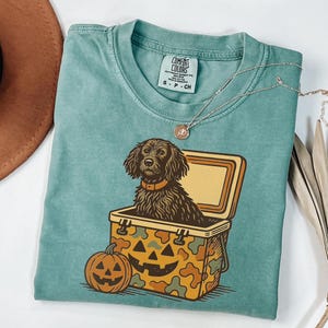 May include: A teal Comfort Colors t-shirt featuring a graphic of a dog inside a cooler with a jack-o'-lantern design. The cooler has a camouflage pattern. A necklace and a brown hat are also visible.