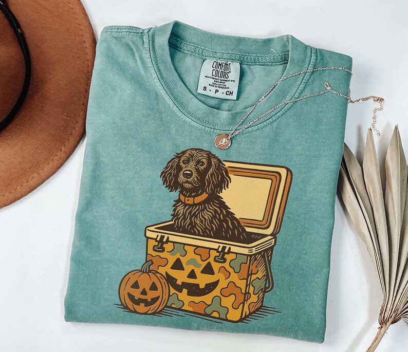 May include: A teal Comfort Colors t-shirt featuring a graphic of a dog inside a cooler with a jack-o'-lantern design. The cooler has a camouflage pattern. A necklace and a brown hat are also visible.