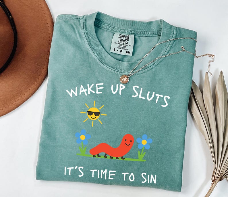 May include: A teal Comfort Colors t-shirt with the text "WAKE UP SLUTS IT'S TIME TO SIN" and a graphic of a smiling sun, worm, and flowers. A silver necklace with a pendant is draped across the shirt.