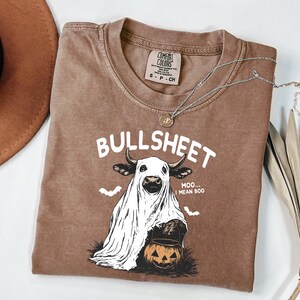 May include: Brown Comfort Colors t-shirt with the word "BULLSHEET" in white. The graphic features a cow in a ghost costume holding a jack-o'-lantern. A silver necklace and a brown hat are also visible.