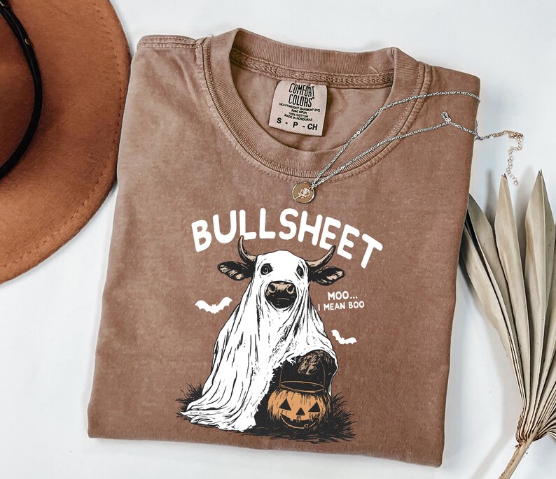 May include: Brown Comfort Colors t-shirt with the word "BULLSHEET" in white. The graphic features a cow in a ghost costume holding a jack-o'-lantern. A silver necklace and a brown hat are also visible.