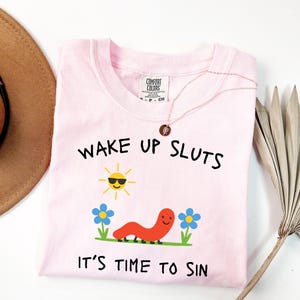 May include: A light pink Comfort Colors t-shirt with the text "WAKE UP SLUTS, IT'S TIME TO SIN". The shirt features a graphic of a smiling sun, a red worm, and blue flowers. A gold necklace is draped on the shirt.