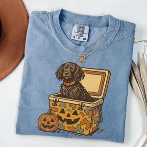 May include: A light blue Comfort Colors t-shirt featuring a Halloween-themed graphic. The graphic includes a black dog in a cooler, a jack-o'-lantern, and a camouflage pattern. A gold necklace is draped across the shirt.