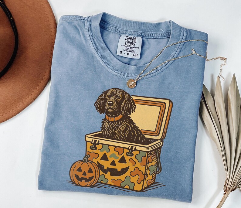 May include: A light blue Comfort Colors t-shirt featuring a Halloween-themed graphic. The graphic includes a black dog in a cooler, a jack-o'-lantern, and a camouflage pattern. A gold necklace is draped across the shirt.