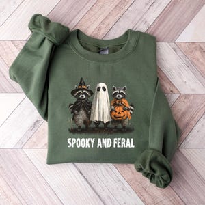May include: Green sweatshirt with a Halloween-themed graphic. The design features a raccoon in a witch hat, a ghost, and a raccoon holding a pumpkin. The text "SPOOKY AND FERAL" is printed below the graphic. The sweatshirt is made of a soft material.