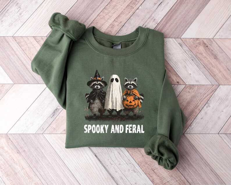 May include: Green sweatshirt with a Halloween-themed graphic. The design features a raccoon in a witch hat, a ghost, and a raccoon holding a pumpkin. The text "SPOOKY AND FERAL" is printed below the graphic. The sweatshirt is made of a soft material.