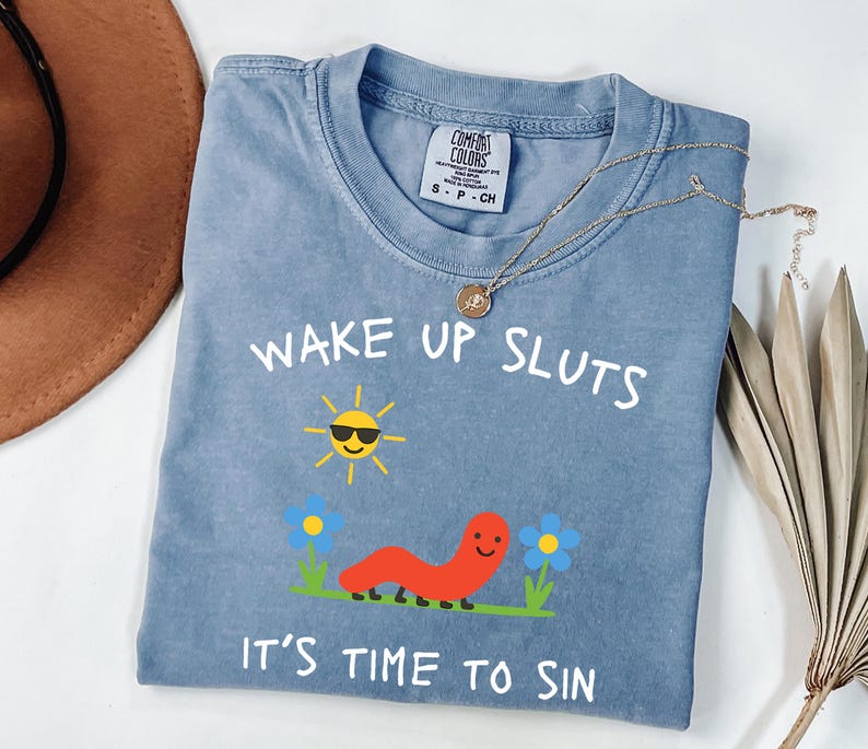 May include: A blue Comfort Colors t-shirt with the text "WAKE UP SLUTS IT'S TIME TO SIN". The graphic includes a smiling sun wearing sunglasses, a red worm, and blue flowers. A gold necklace is draped across the shirt.
