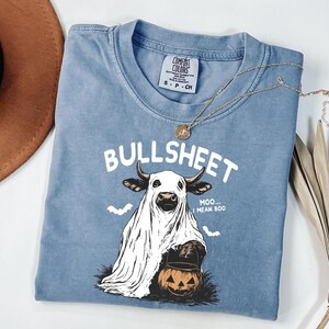 May include: A blue Comfort Colors t-shirt with the word "BULLSHEET" above a cow dressed as a ghost holding a jack-o'-lantern. The shirt also has the text "Moo... I mean Boo" and two bats. A gold necklace is draped across the shirt.