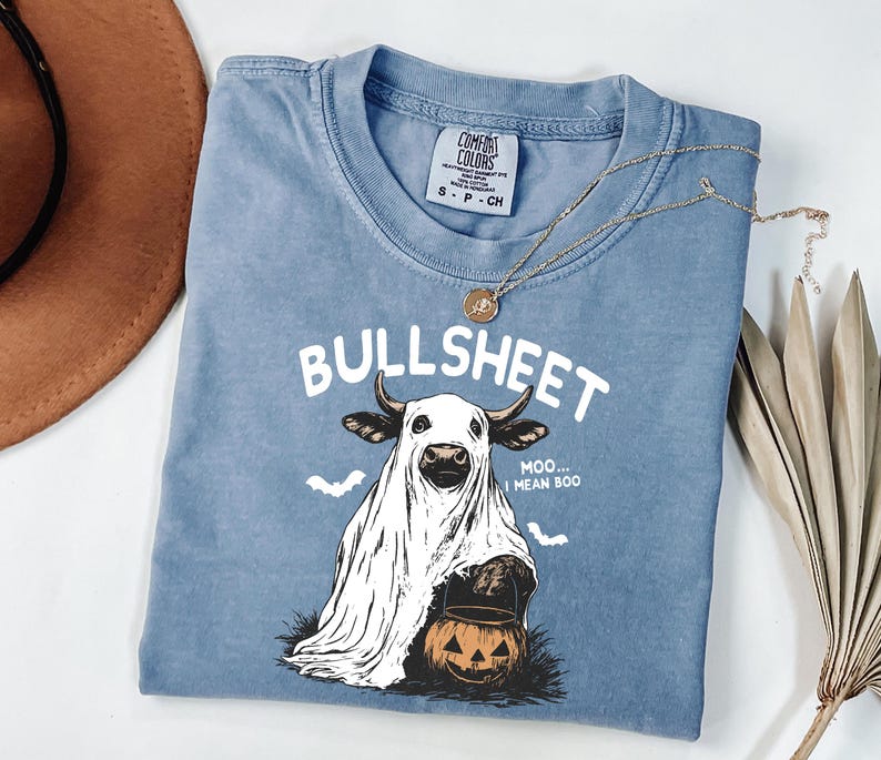 May include: A blue Comfort Colors t-shirt with the word "BULLSHEET" above a cow dressed as a ghost holding a jack-o'-lantern. The shirt also has the text "Moo... I mean Boo" and two bats. A gold necklace is draped across the shirt.